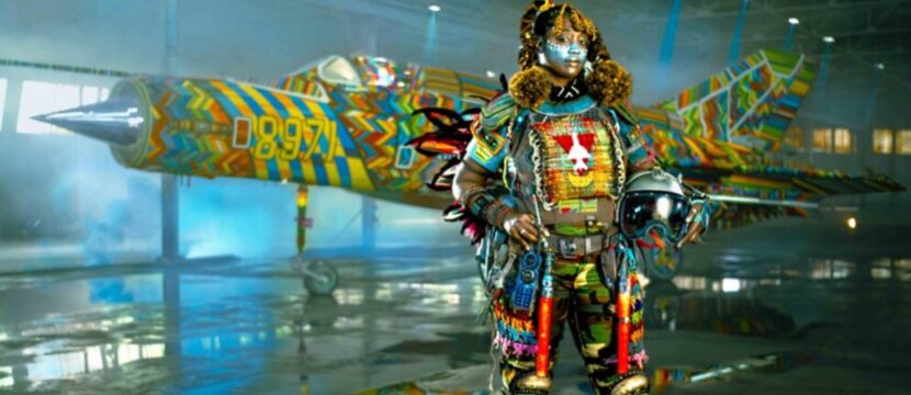 A MiG-21 military flight suit is being modeled by a woman of African descent. She is covered in a brightly colored tribal-inspired armor featuring colorful beads.